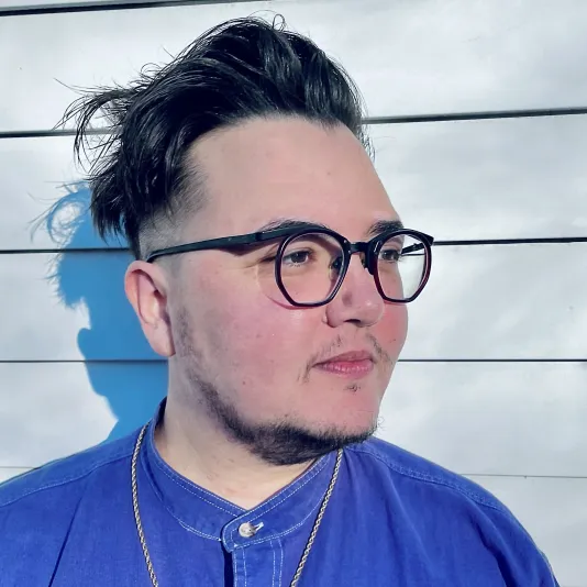 Portrait of Jules Gimbrone looking to the right wearing a blue shirt and black glasses.