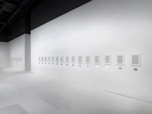 Several rectangular plaster works in white frames are hung horizontally across white gallery walls, each with a horizontal steel plaque below.