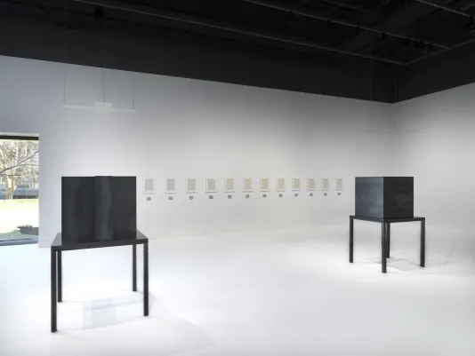 Two dark steel tables and geometric sculptures are surrounded by artworks in white frames hung horizontally in a white walled and floored gallery.