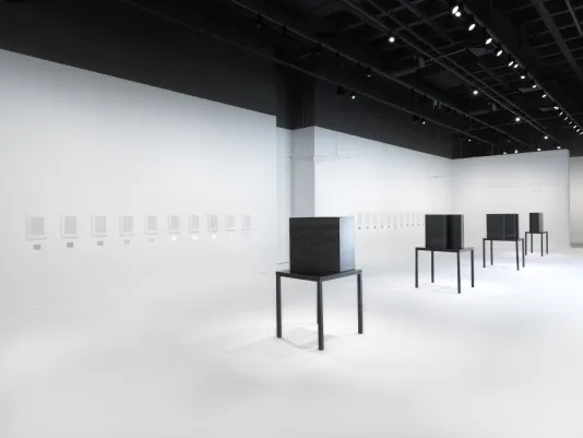 Four dark steel tables and geometric sculptures are surrounded by artworks in white frames hung horizontally in a white walled and floored gallery.