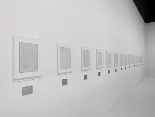 Several page-sized plaster works in white frames are hung horizontally across white gallery walls, each with a small steel plaque below.