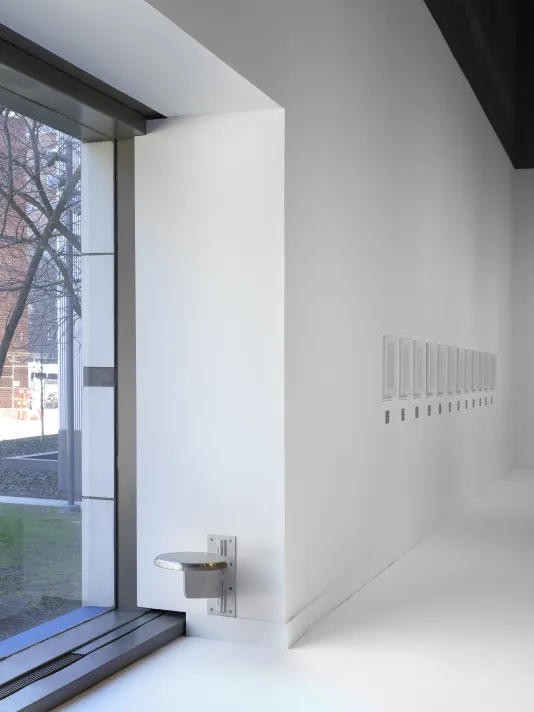 A wall-mounted steel stool is in a white gallery next to a large window.