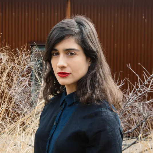Portrait of Naama Tsabar wearing a dark blue sweater and button-down shirt. There are dry branches in the background in front a building.