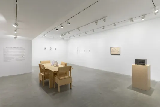 A white walled room with a wooden table and four wooden chairs in the middle of the room. To the right side of the table there is a wooden pedestal with a gray vitrine on top. A row of three glass jars is hung on the left side of a row of eight glass tubes that are hung onto the wall. There is a black framed drawing hung on the wall to the right of the test tubes.