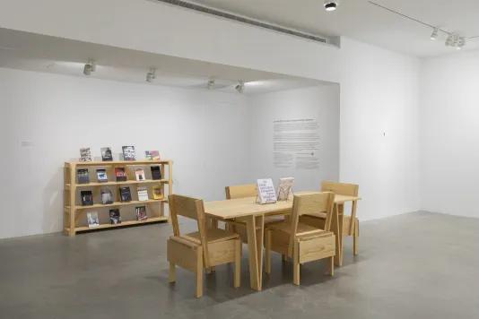 A white walled room with a tan wooden table and four tan wooden chairs in the middle of the room. A wooden bookcase displaying seventeen books is against the wall behind the table.