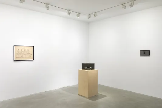 A white walled room with a wooden pedestal with a gray Plexiglas cube on top in the middle. To the left of the pedestal there is a black framed drawing hung on the wall. To the right of the pedestal there is a black and white printed image of Norbert Rilliuex behind a privacy filter with black framing.