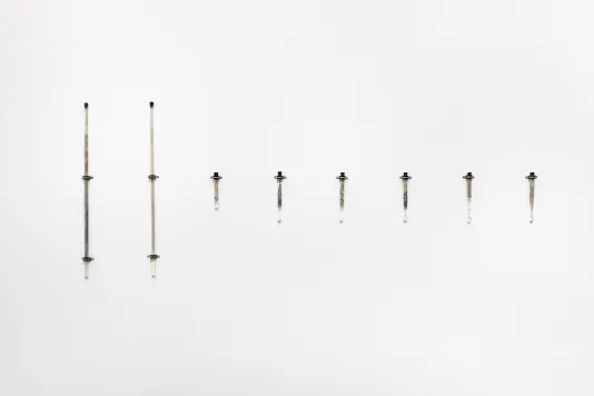 Eight borosilicate glass test tubes are mounted onto a white wall using silver metal brackets. The test tubes all have black stoppers in the top. Reclaimed silver from expired photographic film and paper with sodium hypochlorite can be seen in the test tubes.