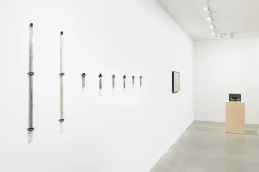 A white walled room with a wooden pedestal and a gray Plexiglas box on top of the pedestal is on the right side of the image. A black framed drawing is hung on the wall to the left of the pedestal. A row of eight glass tubes are hung on to the wall to the left of the drawing.