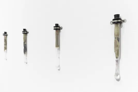 Four borosilicate glass tubes are mounted onto a white wall using silver metal brackets. The tubes all have black stoppers in the top. Reclaimed silver from expired photographic film and paper with sodium hypochlorite can be seen in the test tubes.