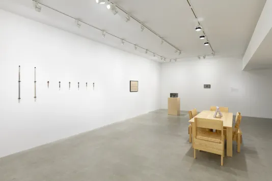 A white walled room with a tan wooden table and four tan wooden chairs in the middle of the room. To the right side of the table there is a wooden pedestal with a gray Plexiglas cube on top. There is a black framed drawing hung on the wall to the left of the pedestal. A row of eight test tubes are hung onto the wall to the left of the drawing.