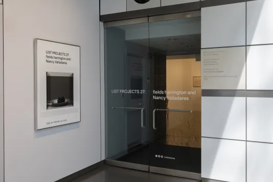 A white poster that reads “LIST PROJECTS 27: fields harrington and nancy Valladares” with an image of a gray Plexiglas cube is hung next to two glass gallery doors with the same text and beige glowing light.