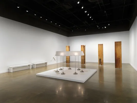 In the foreground, seven standing lamps are conjoined by a single ring-shaped lampshade. This large sculpture sits on a low pedestal. Behind it, four oblong wooden sculptures hang. A smaller chevron-shaped white sculpture is affixed to a wall in the background.