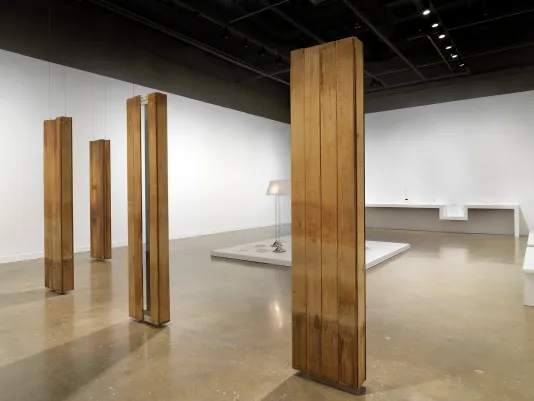 In the foreground, four oblong sculptures hang in formation, each made from planks of reclaimed wood. A standing lamp peaks out from behind one of the sculptures. An uneven shelf runs along the back wall.