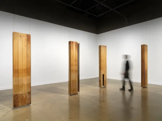 Four oblong sculptures hang in formation, each made from planks of reclaimed wood. They hover an inch or so above the floor. Some sections of wood are missing and replaced with glass. A person walks among the sculptures.