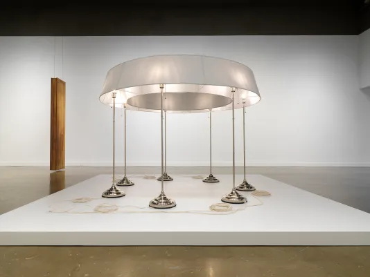 On a low pedestal in the foreground, seven standing lamps are conjoined by a single ring-shaped lampshade. The photograph is taken from a low angle so the “hole” in the center of the lampshade is visible. A neat coil of pale cord sits near the base of each lamp. An oblong wooden sculpture hangs in the background, monolith-like.