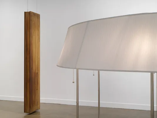On the right, an unusually large lampshade is partially visible and illuminated from within. Three lamp stems, each with a metal pull chain, extend downwards from different parts of the shade. On the right, a sculpture made from reclaimed wooden planks hangs monolith-like, hovering an inch or so above the floor.