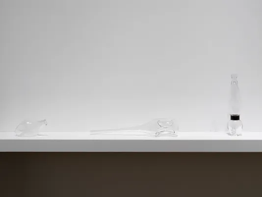 Three glass sculptures sit on a shelf affixed to a white wall. They resemble odd vessels or bottles, with rounded indentations or craters. The vessel in the middle has a long and subtly tapering neck, and sits horizontally. The one on the right sits upright, and appears to be choked by a tight band of metal near its base.
