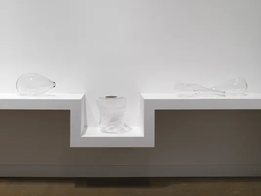  Four glass sculptures sit on a shelf against a white wall. They resemble stretched, deformed, or indented vessels. The shelf sinks to accommodate a bulkier sculpture, the top of which is level with the rest of the shelf. Its flat top contains a round drain cover, like one you would find in a shower.