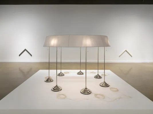 On a low pedestal in the foreground, seven standing lamps are conjoined by a single ring-shaped lampshade. A neat coil of pale cord sits near the base of each lamp. The sculpture is flanked, in the background, by two chevron-shaped wall pieces. The one on the left is dark, while the one on the right is pale.