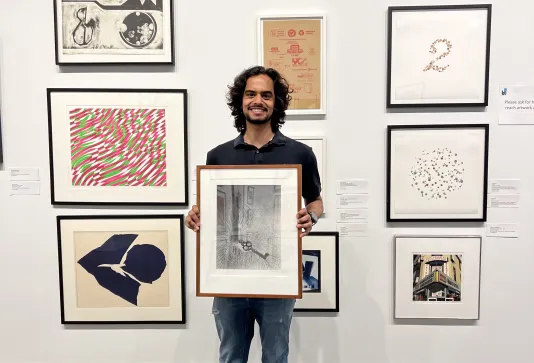 A student with brown hair holds a large framed artwork in front of a white gallery wall with numerous other framed works of art.