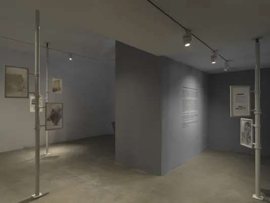 A thick wall with text on it separates a gallery into two sections. The edge of a wooden chair peeks out from behind its left side. On either side, metal poles extend from floor to ceiling. Framed works on paper with images and text are attached to each pole. The frames appear to be able to rotate around the poles.