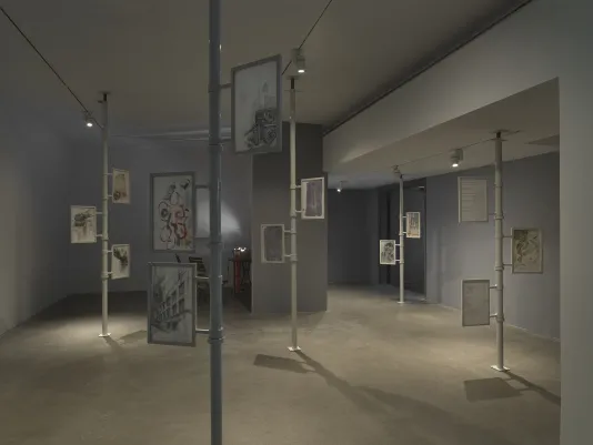 Five metal poles extend from floor to ceiling in a gallery. Two or three framed works on paper are attached to each one, including outlines of plants, drawings of plants, more abstract compositions, and lines of text. In the background on the left, a projector on a table casts an abstract light pattern across the wall, accompanied by two chairs.