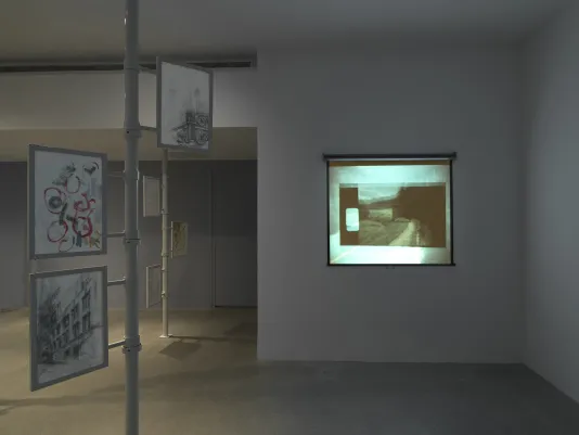 On the right, a film is projected onto a pull-down screen against a wall. An image of a car driving on a curved road is overlaid with another smaller, less discernible image with a sprocket hole. On the left, two metal poles extend from floor to ceiling, each with three framed works of paper attached. The two drawings closest to the camera show the facade of a multi-story building and a more abstract composition with squiggly circles.