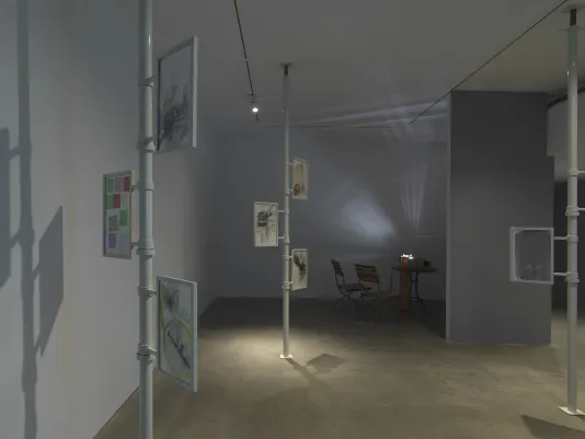 Three metal poles extend from floor to ceiling in a gallery. Two or three framed works on paper with layered compositions are attached to each one. The frames appear to be able to rotate around the poles. In the background on the left, a projector on a table casts an abstract light pattern across the wall, accompanied by two chairs.