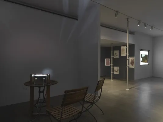 A makeshift projector sits on a wooden table, accompanied by two matching chairs. Two cords and a strip of material are attached to the device, which projects a faint circle of light onto the back wall. On the right and in the background, two floor-to-ceiling poles with drawings attached and a rectangular, more conventional projected image are visible.