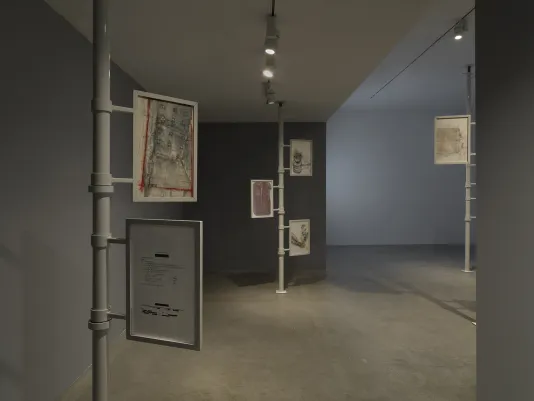 Three metal poles extend from floor to ceiling. Two or three framed works on paper are attached to each one. Nearest to the camera, one work appears to be a printed document with some lines redacted; another is a drawing with a geometric layout resembling an architectural plan. The frames appear to be able to rotate around the poles.