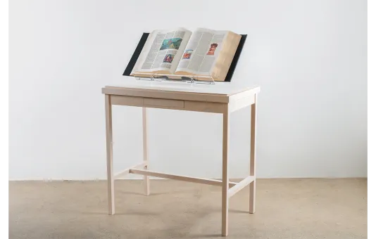 Image of a light cream wooden table displaying an open encyclopedia book within in a white walled room.
