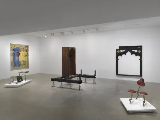 A white gallery with five artworks on display. On the left wall hangs a gold tapestry depicting two figures, one standing and one kneeling. Two small creature-like sculptures made of metal and fabric sit on low pedestals. In the center of the room is a vertical wooden bathtub sculpture on metal supports. A large black frame-like wooden sculpture hangs on the back wall.
