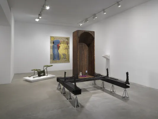 In the foreground, a vertical wooden bathtub sits on horizontal wooden beams, which are held up by eight metal supports. Inside the bathtub are small painted scenes and a red and blue checkerboard pattern on the base. In the background, a gold tapestry depicting two figures hangs. On the floor to the left, a four-legged creature-like sculpture sits on a low pedestal. On the right wall is a small white shelf with a plexiglass case.