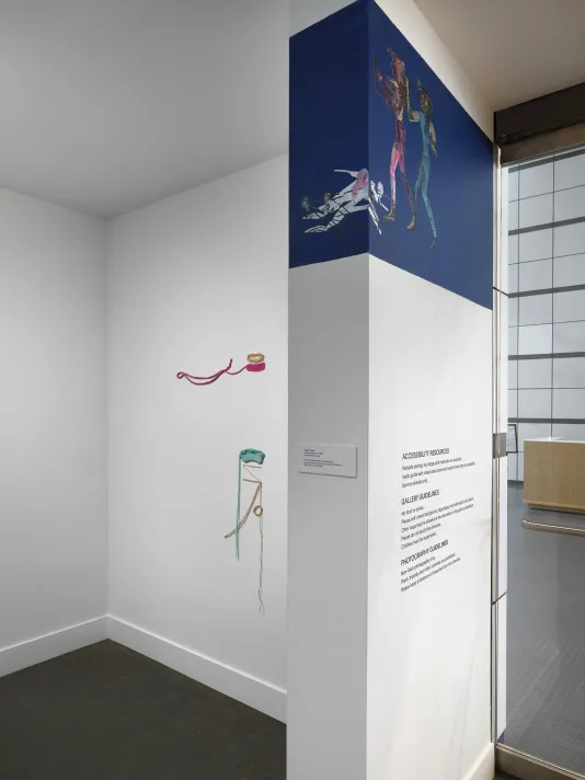 A corner, wrap-around, wall of the entrance to a gallery space. On the top of the wall in the foreground, a painting depicting colorful Egyptian figures on a blue background wraps around the corner. In the background, two abstract, string-like, fiber artworks are on display stacked on top of each other. The topmost piece is a pink-like color, the below piece is a blue-green color.