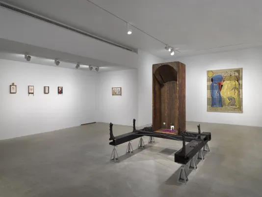 In the foreground, a vertical wooden bathtub sits on horizontal wooden beams, which are held up by eight metal supports. Inside the bathtub are small painted scenes and a red and blue checkerboard pattern on the base. On the left walls, five small drawings hang in wooden frames. In the rear, a golden tapestry depicts two figures, one standing and one kneeling.