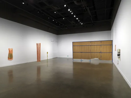 A white gallery space with cement floor displaying several artworks scattered throughout the space. On the left side, two orange net-like fiber sculptures hang, one off the wall and the other from the ceiling. On the right, a large, brown, rectangular tapestry with blue gridded lines hangs from the ceiling. Smaller artworks are displayed in the background and on the right wall.