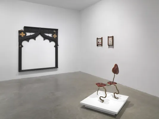 A white gallery with four artworks on display. On the left a large black frame-like wooden sculpture hangs on the back wall. To the right of the frame-like sculpture, two small drawings hang in wooden frames. In the foreground, a small creature-like sculpture made of metal and fabric sits on a low pedestal.