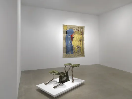 In the foreground, a small creature-like sculpture made of metal and fabric sits on a low pedestal. A small piece of fabric showing an animal hangs from the central axis or “spine” of the sculpture. In the background hangs a gold tapestry depicting two figures, a standing blue one and a cartoon-like kneeling one.