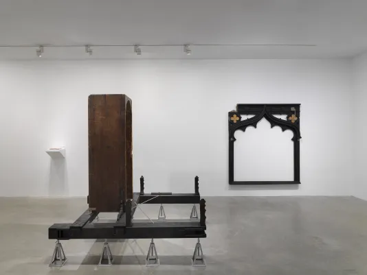 In the foreground, a vertical wooden bathtub sits on horizontal wooden beams, which are held up by eight metal supports. In the background on the left side is a small white shelf with a plexiglass case. On the right hangs a large black frame-like wooden sculpture.