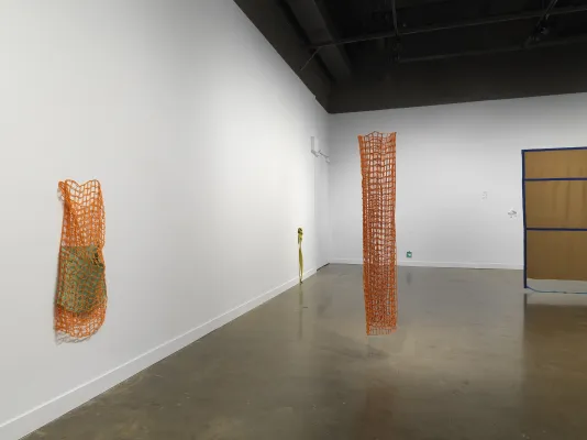 On the left, an orange net-like fiber sculpture hangs off the wall encasing a smaller, green fiber cylindrical piece. Suspended from the ceiling in the center of the image, another orange net cylinder is suspended from the ceiling. Several smaller fiber sculptures are affixed to the wall in the background. In the right of the frame, a large brown tapestry peeks into the frame.