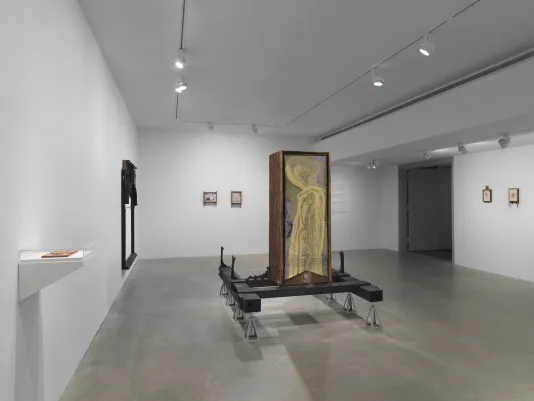In the center of a gallery, a golden abstract tapestry hangs on the back of a wooden bathtub sculpture that is raised on black planks and metal supports. On the left wall in the foreground is a small white shelf with a plexiglass case. Further along the wall hangs a large black frame-like wooden sculpture. Across the background two small drawings in wooden frames hang on the left and two other drawings in wooden frames hang to the right of an entryway.