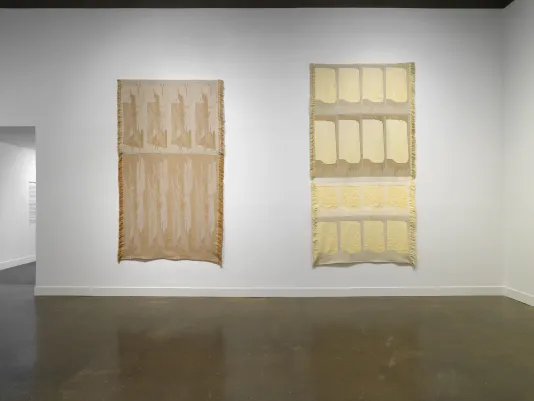 Two tapestries, similar in scale and color, hang side by side. Each depicts abstracted forms that are repeated in a grid with slight variations, as if some rows were stretched. They are brown, beige, yellow, and gold.