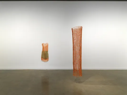 A white gallery with two artworks on display. On the left, a woven orange net-like sculpture hangs on the wall encasing a smaller, green fiber sculpture. On the right, a similar sculpture (though taller and without the inner green form) is suspended from the ceiling.