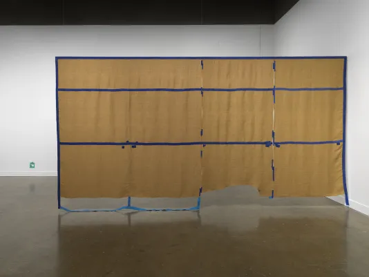 A large rectangular brown tapestry hangs from the ceiling of a gallery. Irregular strips of tape-like blue textile create a grid on top of the brown, with some of the blue strips dangling on the floor. In the background on the left, a tiny green fiber artwork is affixed to the wall close to the floor.