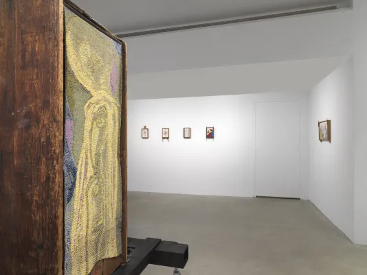 On the left, a golden abstract tapestry hangs on the back of a wooden bathtub sculpture that is raised on black planks and metal supports. The tapestry has swirling forms of yellow and gold, with some sections of blue near the periphery. In the background, five small drawings hang in wooden frames on a white wall.