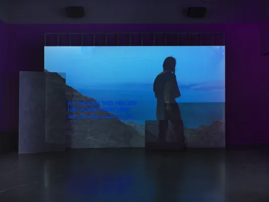 A deep blue video depicting a person looking out from a mountain onto a body of water is projected onto an uneven surface of overlapping rectangular panels. Overlaid on the video footage, English and Arabic text reads “As though this melody has always been here”.