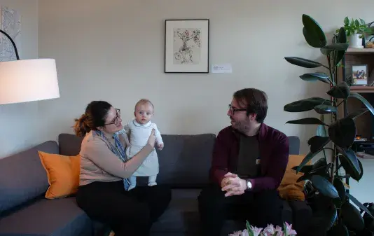 A man and woman holding a baby are sitting on a gray couch with a framed artwork on the wall behind.