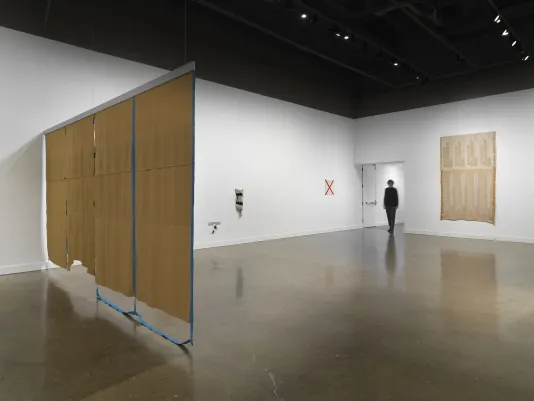 In the foreground on the left, a large brown tapestry is suspended from the ceiling. Blue strips of textile grid the tapestry, with some of the tape-like strips dangling onto the floor. On the right, a smaller gold-beige tapestry hangs on the wall. On the back wall, three smaller fiber artworks hang on the wall, including a small off-white square with a red X hung next to the entrance to the space. A blurred silhouette of a person stands in the entryway.