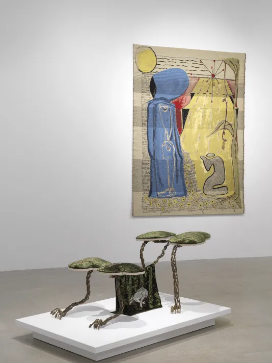 A white gallery with two artworks on display. In the foreground, a small creature-like sculpture made of metal and fabric sits on a low pedestal. In the background hangs a gold tapestry depicting two figures, one standing and one kneeling.