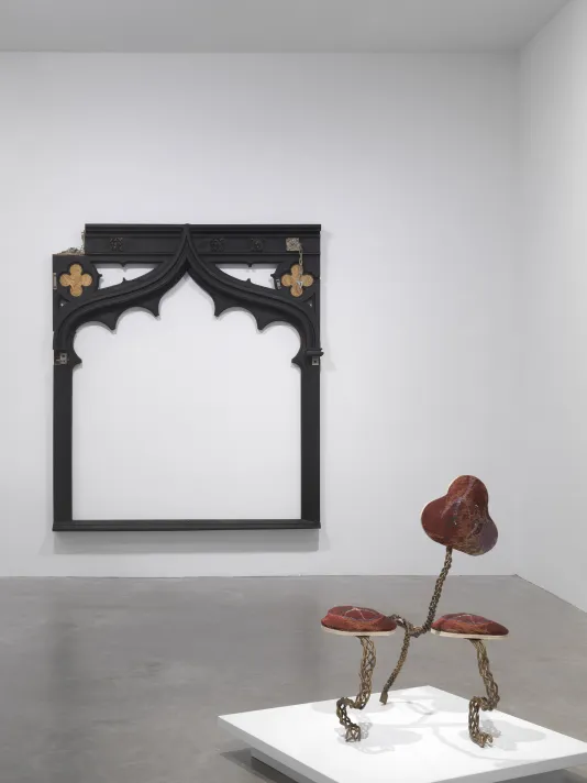 A white gallery with two artworks on display. In the background, a large black frame-like wooden sculpture hangs on the wall. In the foreground, a small creature-like sculptures made of metal and fabric sits on a low pedestal.